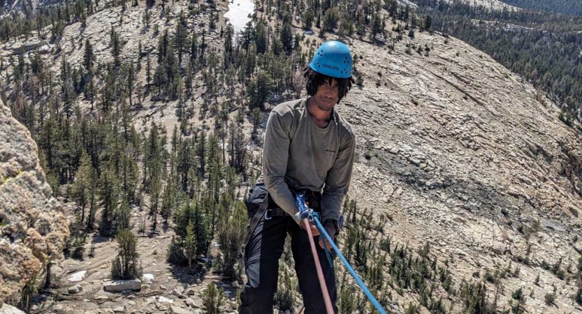 A person wearing safety gear is secured by ropes as they stand near the edge of a cliff, high above a mountainous landscape. 